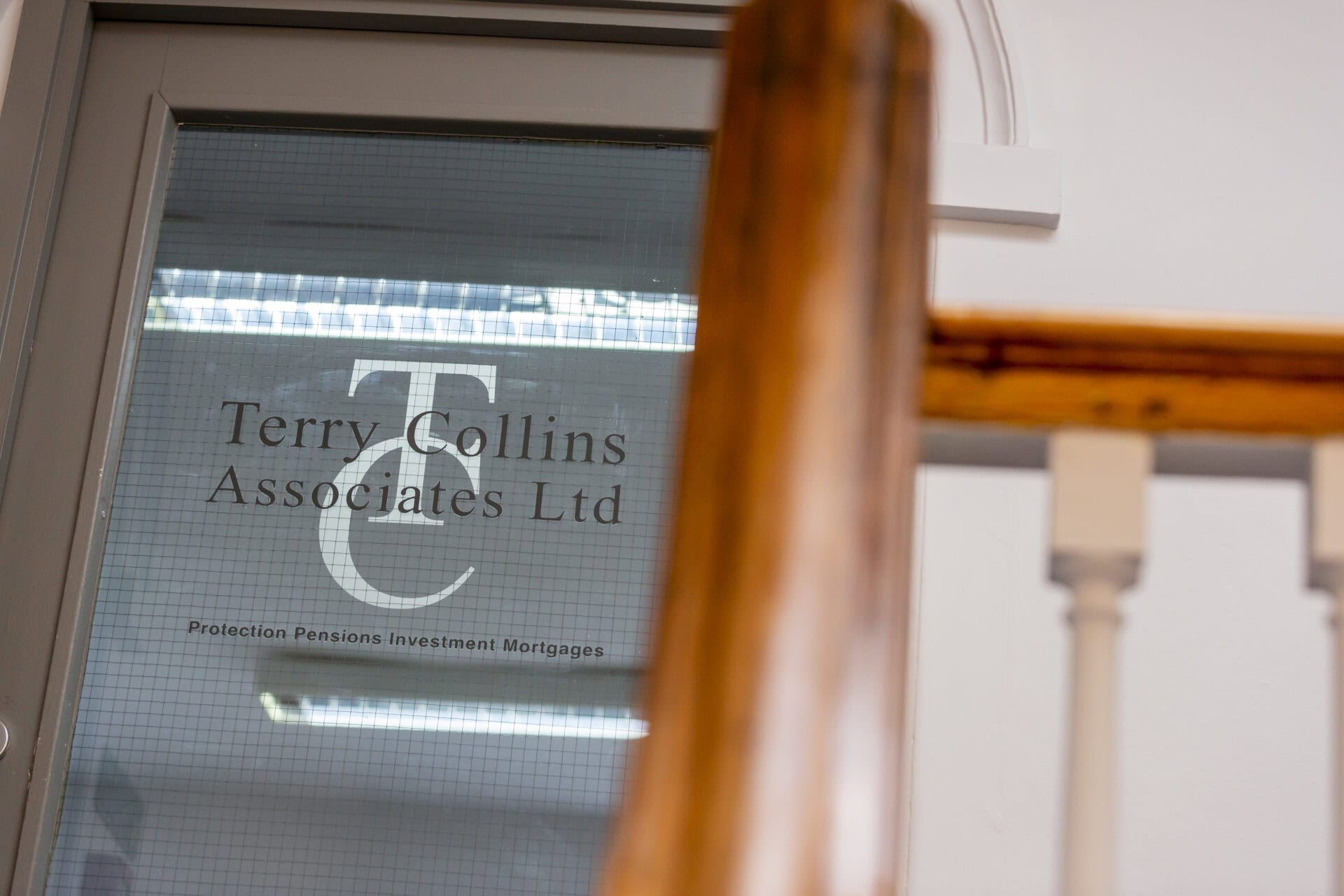 Office door with "Terry Collins Associates Ltd" logo, offering services in protection, pensions, investment, and mortgages. View partially obscured by a wooden staircase railing.