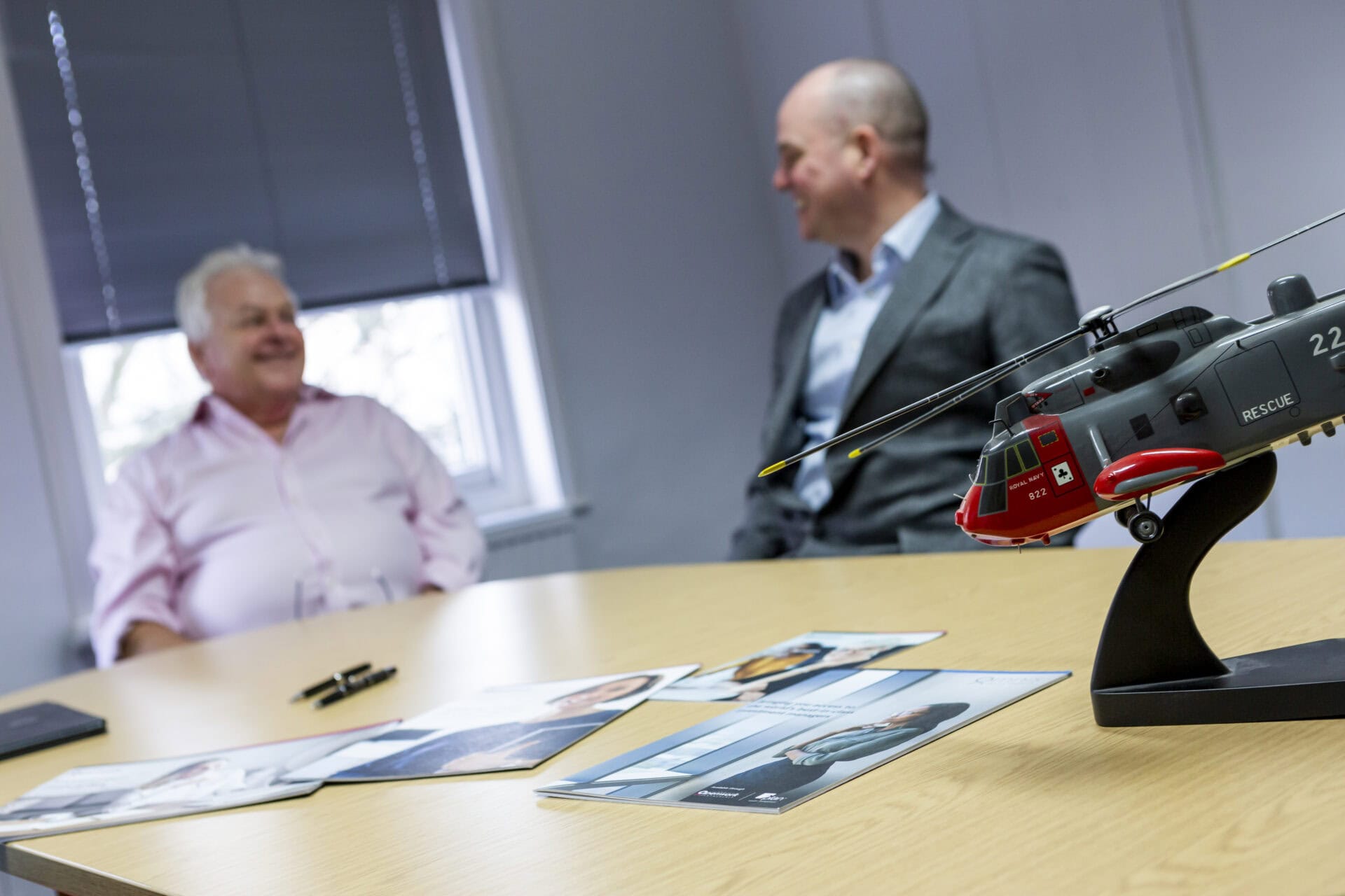 Two men seated at a table with brochures and a model helicopter in the foreground.