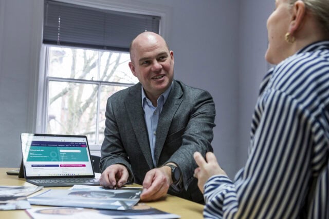 A man in a suit and a woman in a striped shirt discuss papers at a table with a laptop showing a website. A window in the background reveals tree branches.